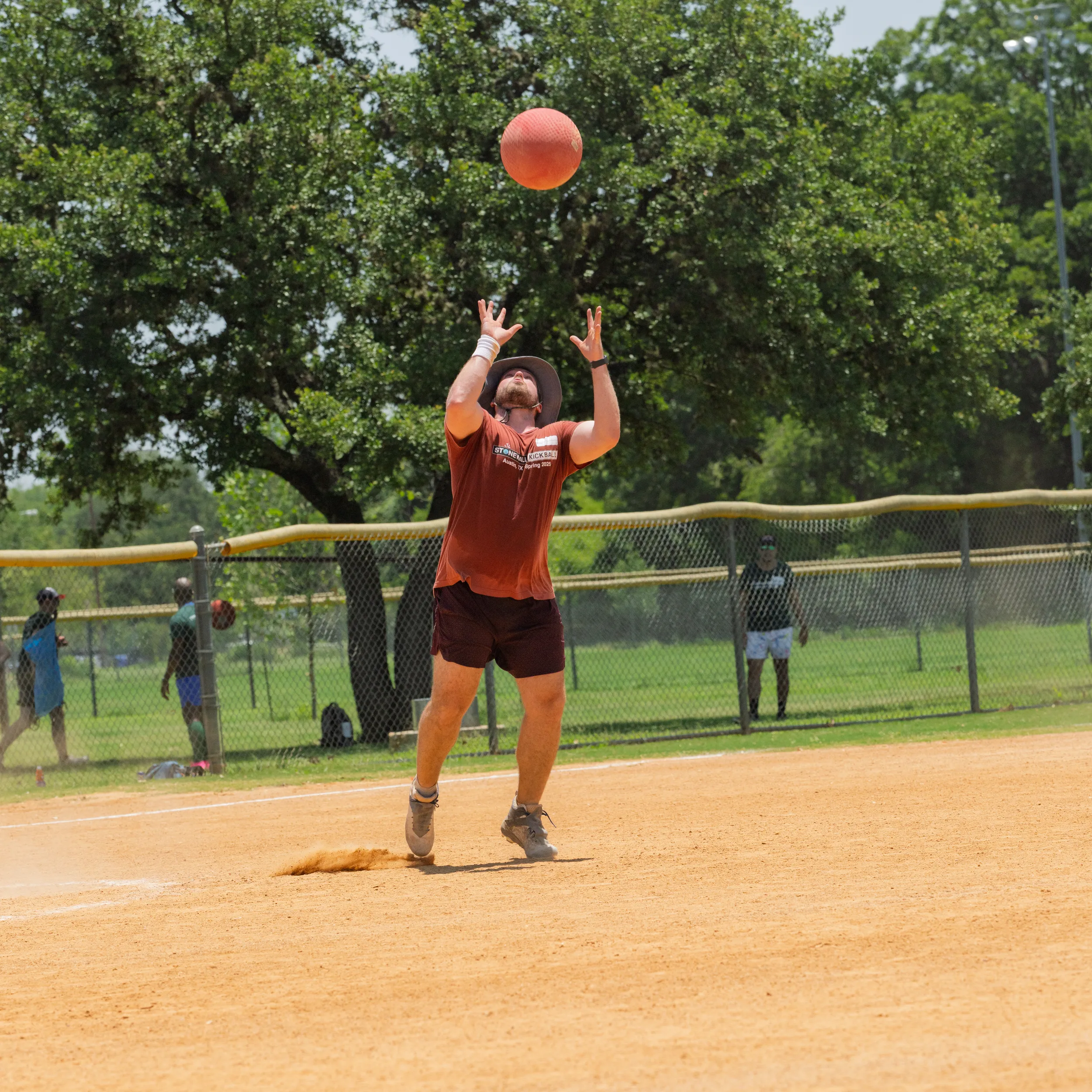 On a sunny kickball field, a player in a maroon T-shirt and shorts, athletic shoes, and a wide-brim hat stands on the infield with arms raised to catch a red kickball dropping overhead, dust kicking up at their feet. A chain-link fence, trees, and a few other players watching from the outfield frame the scene.