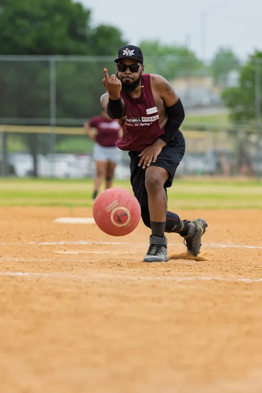 A player wearing a maroon Stonewall Kickball shirt, black shorts, knee brace, sunglasses, and a cap is in the process of pitching a red kickball on a dirt field, with another person visible in the background.