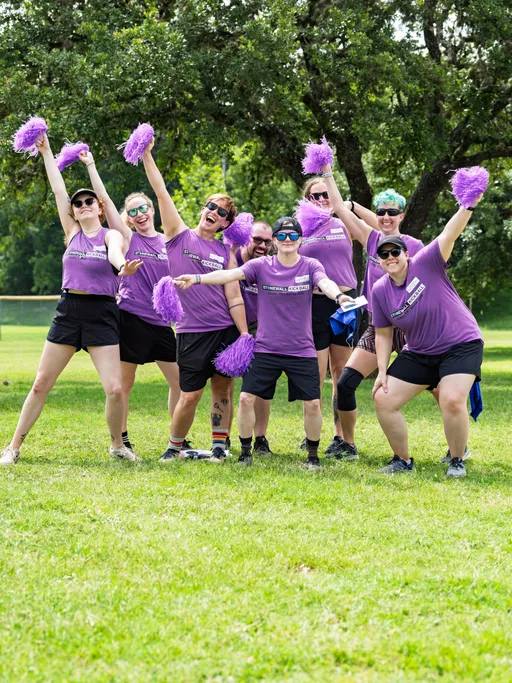 A group of players wearing matching purple "Stonewall Kickball" shirts, black shorts, and sunglasses pose energetically on a grassy kickball field, holding purple pom-poms and smiling with arms raised.
