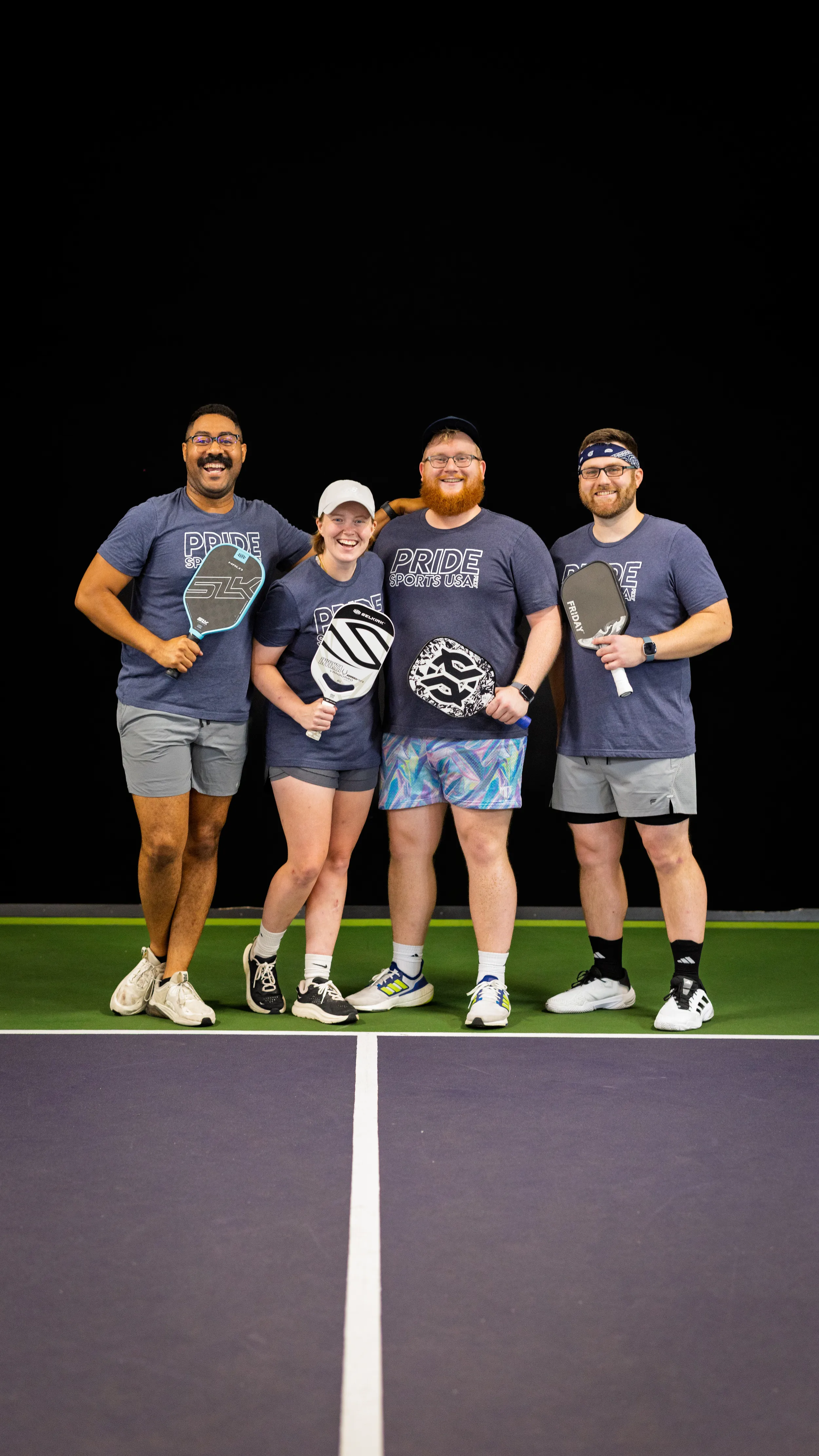 Four pickleball players pose arm-in-arm on a green-and-purple court, holding patterned paddles and wearing matching “PRIDE SPORTS USA” shirts, shorts, and athletic shoes; one has a cap and another a headband. The group smiles against a dark backdrop, capturing a team moment from the Pride Pickleball Summer 2025 season.