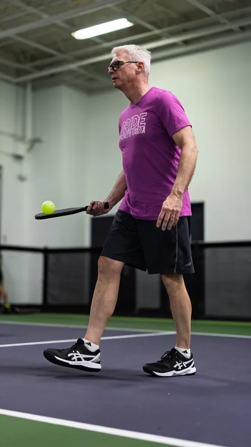 A pickleball player wearing a purple "Pride Sports USA" t-shirt, black shorts, and athletic shoes is walking across an indoor court, holding a paddle and preparing to hit a pickleball.