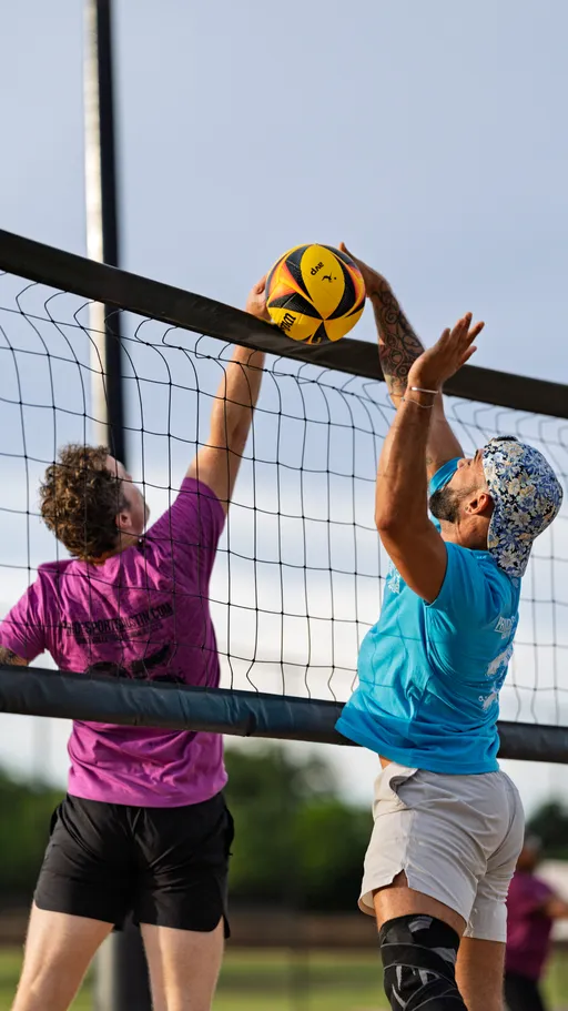 Two volleyball players jump at the net, both reaching for the ball above the net; one player wears a purple shirt and black shorts, and the other wears a blue shirt, light shorts, floral hat, and a knee brace. They are competing on an outdoor sand volleyball court with green trees in the blurred background.