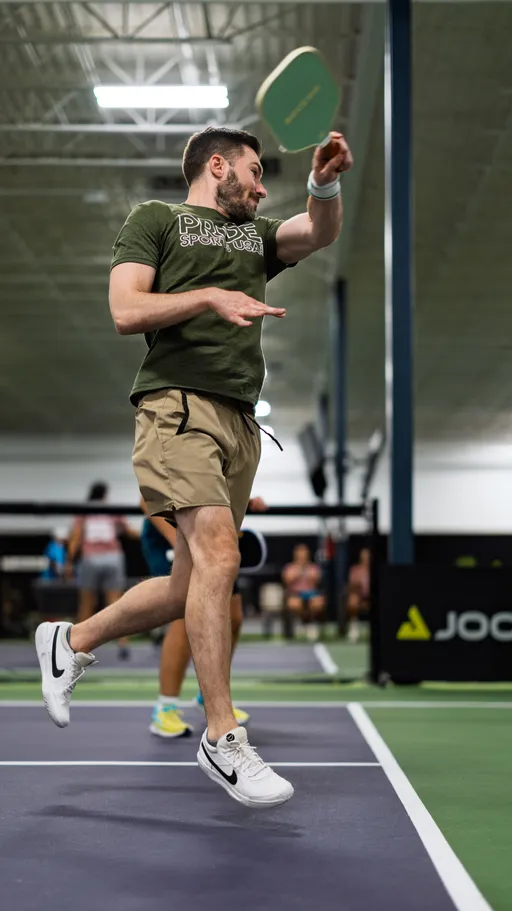 A player in a "Pride Sports USA" shirt and athletic shorts is mid-jump, swinging a pickleball paddle on an indoor pickleball court, with other players and spectators visible in the background.