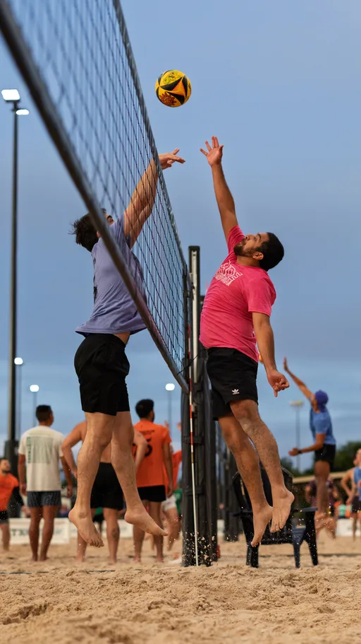 Two sand volleyball players are jumping at the net, reaching for the ball mid-air; one wears a purple shirt and black shorts, while the other wears a pink shirt and black shorts. The game takes place outdoors on a sandy court surrounded by other players and sporting equipment under evening lights.