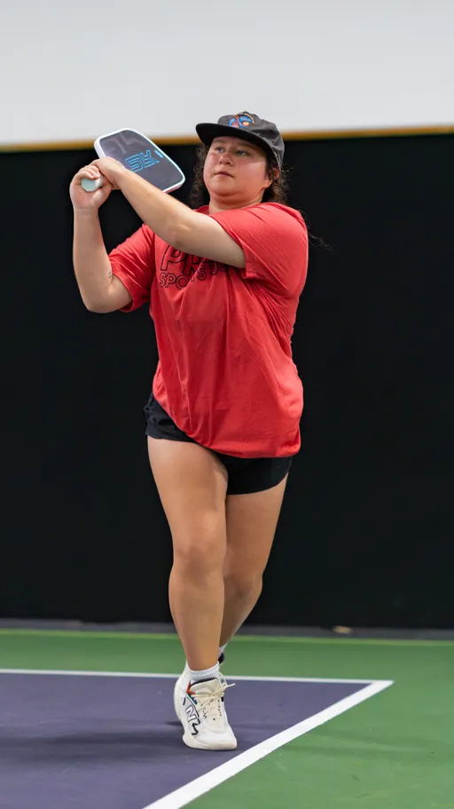 A pickleball player in a red shirt, black shorts, and white sneakers is preparing to hit a shot on an indoor court, holding a paddle mid-swing and wearing a black cap.