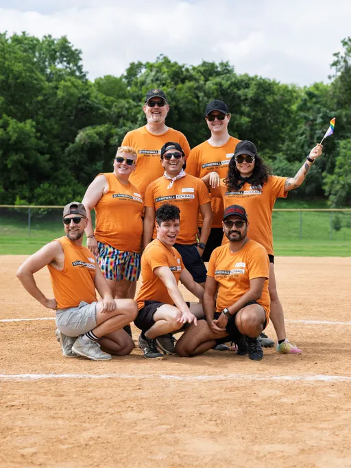 A group of kickball players wearing matching orange "Stonewall Kickball" shirts pose together on a kickball field, with some kneeling in front and others standing behind; one person is holding a small rainbow flag, and the background shows a grassy, tree-lined area.