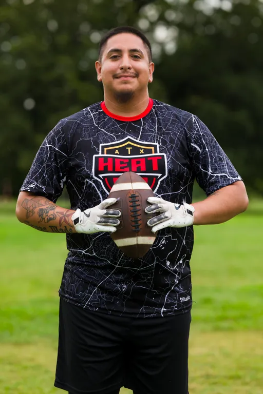 ATX Heat player stands on a grassy field during Gay Bowl 2025 practice, holding a football with both hands at chest level. The athlete wears white receiver gloves, black shorts, and a black jersey with a lightning pattern and a red-and-gold “ATX HEAT” crest, with trees blurred in the background.