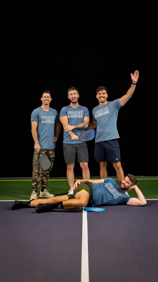 Four pickleball players pose together on a court, all wearing blue "Pride Pickleball" shirts; three stand holding paddles while one reclines playfully on the court in front, with a relaxed and cheerful atmosphere.