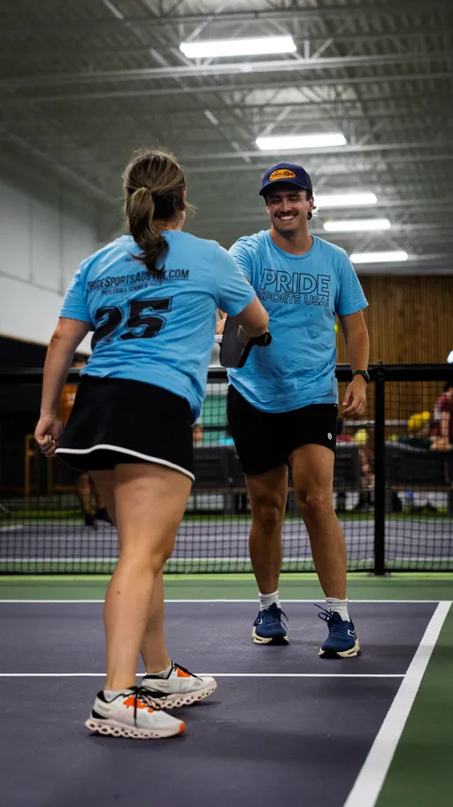Two pickleball players wearing matching blue "Pride Sports USA" shirts are exchanging a high-five on an indoor court, with paddles in hand and a net in the background.