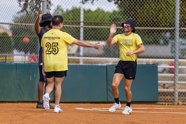 On a kickball field, three Pitch Please players in matching yellow PRIDE shirts celebrate at home plate with a high-five, one jersey showing the number 25 and “pridesportsaustin.com.” A chain-link backstop with green padding and bleachers frames the scene, a bright orange kickball is blurred in the background, and some players wear bucket hats, sunglasses, and athletic shoes.