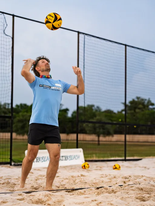 A volleyball player wearing a light blue Stonewall league shirt and black shorts is preparing to serve a volleyball on a sand court, with other volleyballs and a net visible in the background.
