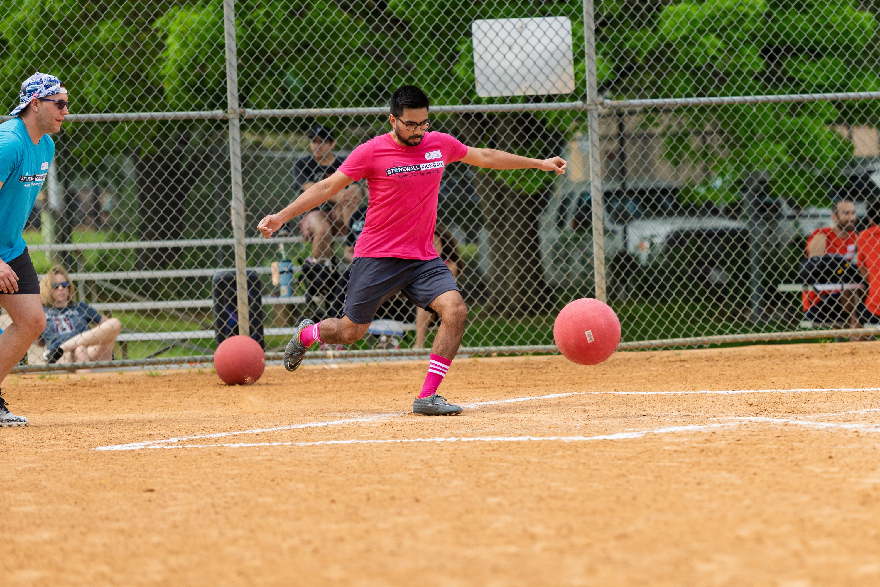 A player in a pink shirt, dark shorts, and pink striped socks is kicking a red kickball on a dirt field while another player in a blue shirt watches nearby; the scene takes place outdoors with a chain-link fence and spectators in the background.