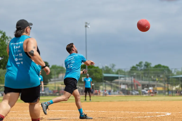 Two kickball players in blue shirts and athletic shorts are on a field; one player has just kicked a red kickball high into the air while the other runs forward. The scene takes place on an outdoor kickball field with green trees and a fence in the background.