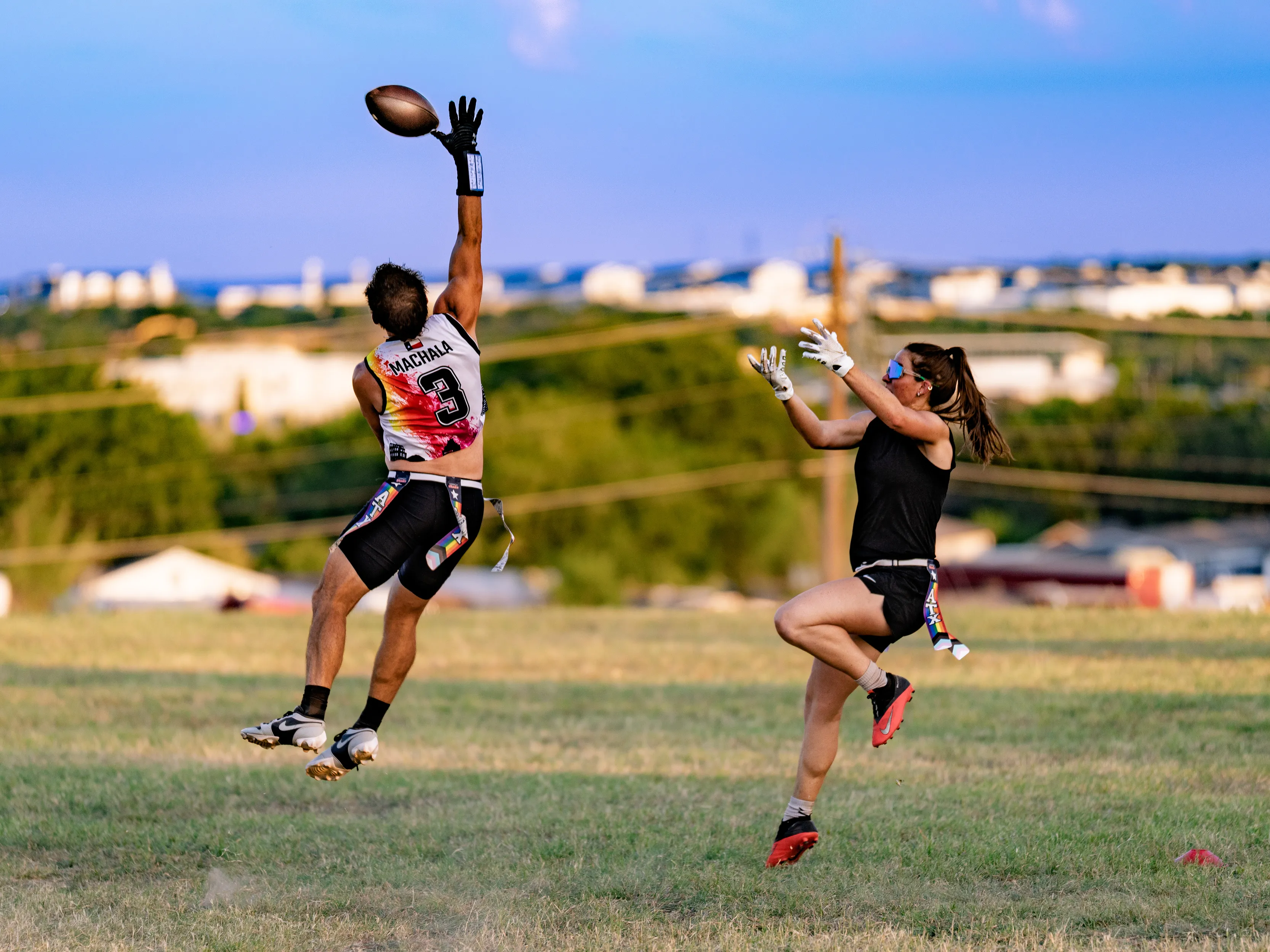 On a grassy practice field, an ATX Heat (Black) receiver in a white-and-black No. 3 jersey, gloves, and a flag belt leaps with one arm extended to snag a high pass while a defender in black athletic gear, gloves, sunglasses, and bright cleats jumps to contest. Cones mark the drill area with a cityscape in the distance under a clear sky.