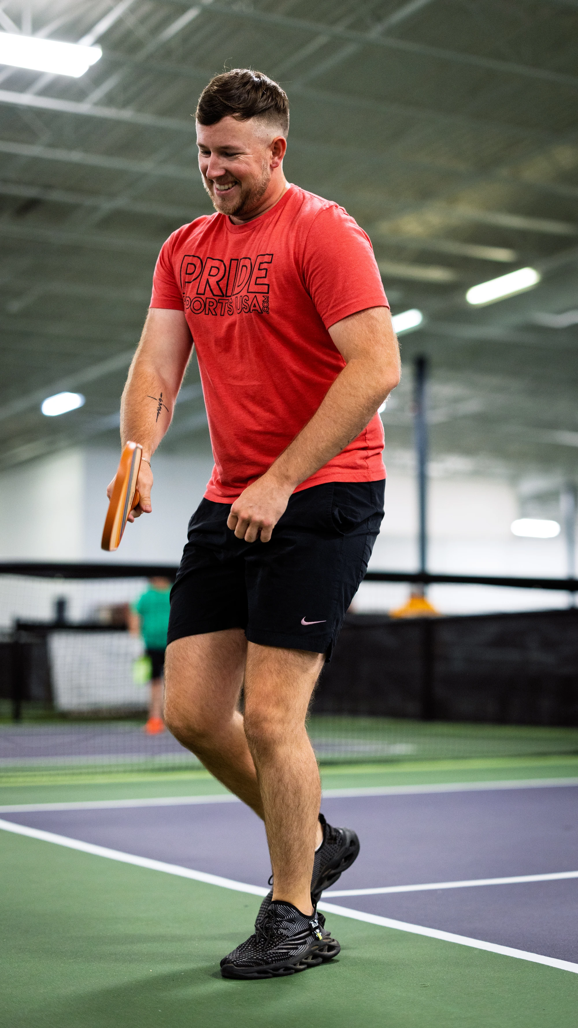 A pickleball player in a red "Pride Sports USA" shirt and black shorts is actively moving on an indoor pickleball court, holding a paddle and appearing focused on the game. The setting is a well-lit sports facility with other players and courts visible in the background.