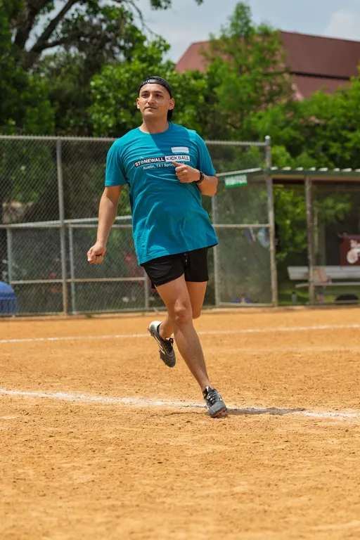 A player wearing a teal "Stonewall Kickball" t-shirt, black shorts, and a backwards cap is actively running on a dirt kickball field, with a chain-link fence and trees in the background.