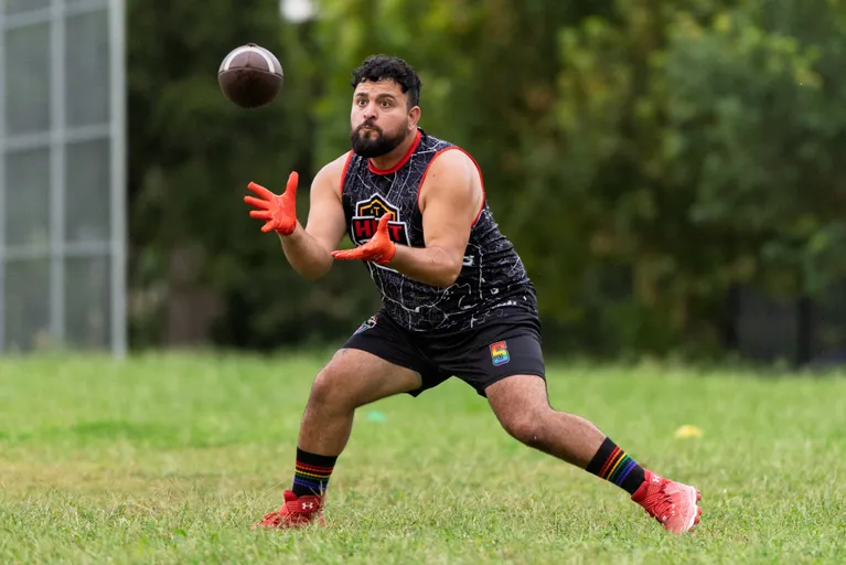 An ATX Heat (Orange) player crouches on a grassy field, hands raised in orange gloves to catch a football in midair during practice for Gay Bowl 2025. The athlete wears a black sleeveless team jersey, black shorts with a pride patch, rainbow-striped socks, and cleats; trees and fencing blur in the background.