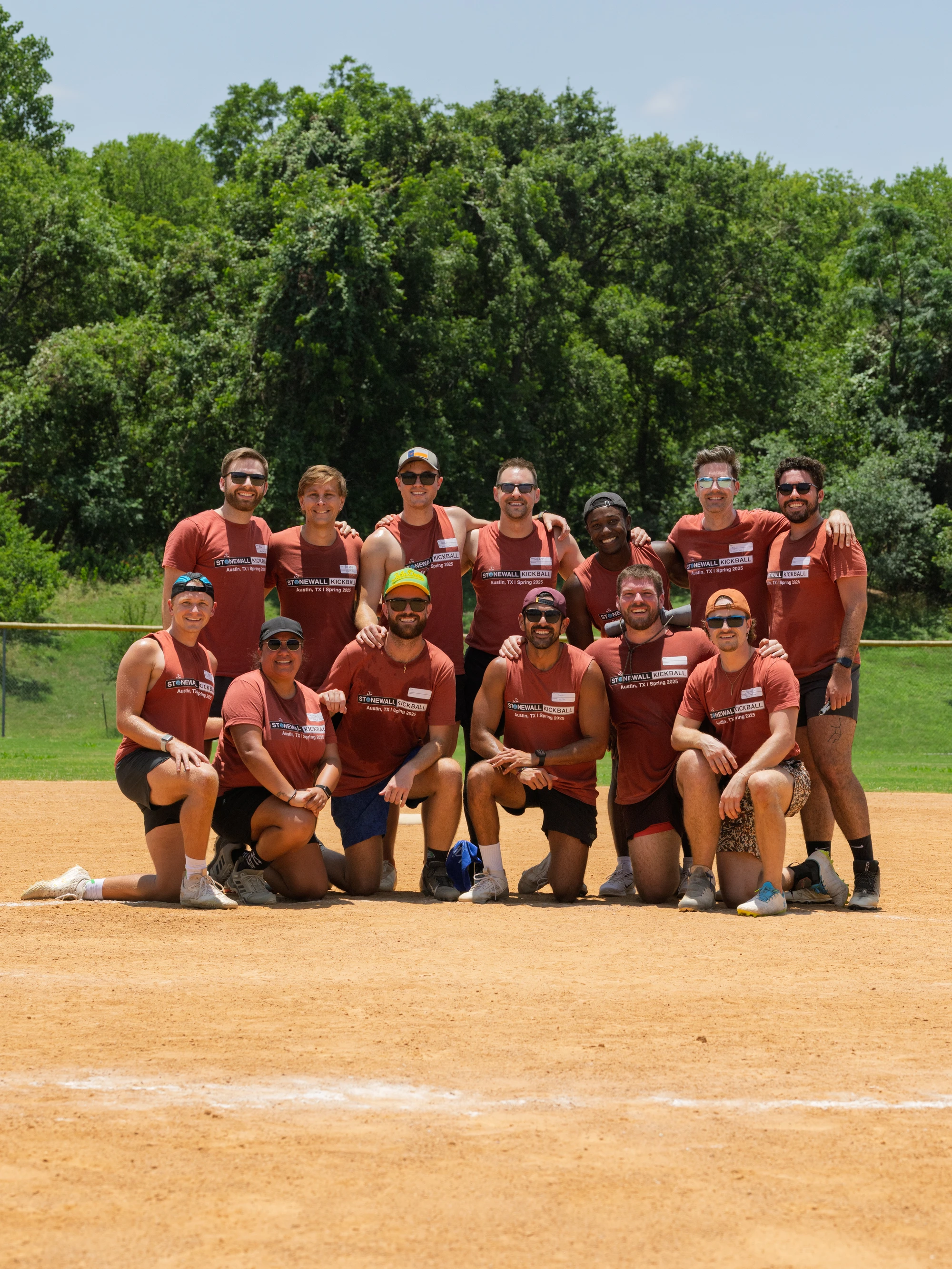 A group of kickball players pose together on a dirt field, all wearing matching red t-shirts, shorts, and various athletic shoes; some have sunglasses, hats, or headbands, with trees and green grass visible in the background.
