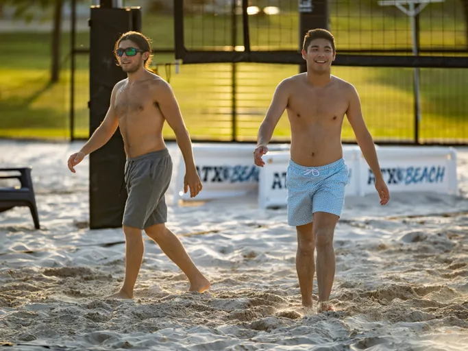 Two volleyball players walk across a sandy court, both wearing swim trunks; one player also wears sunglasses. The net and nearby banners are visible in the background, with sunlight and greenery surrounding the outdoor setting.