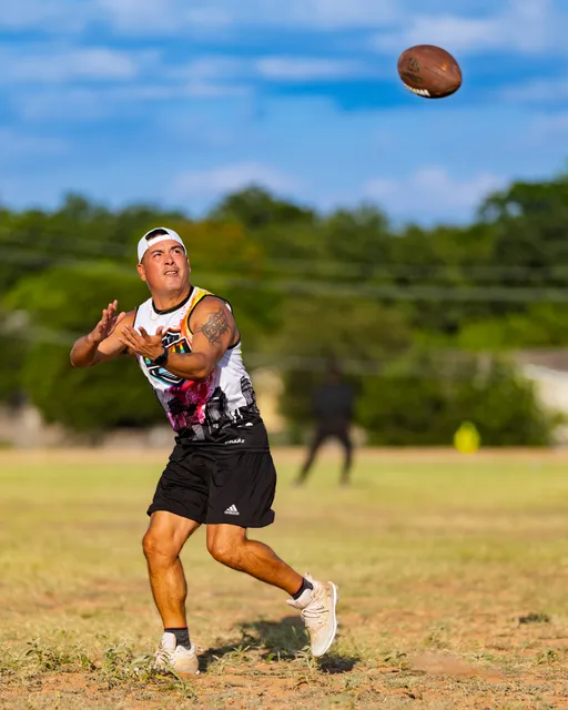 During Gay Bowl 2025 practice, an ATX Heat (Black) player tracks a football in midair, hands up to catch while striding across a dry grass field. The athlete wears a white backward cap, a colorful ATX Heat sleeveless jersey, black shorts, and white cleats, with a blurred teammate and trees in the sunny background.