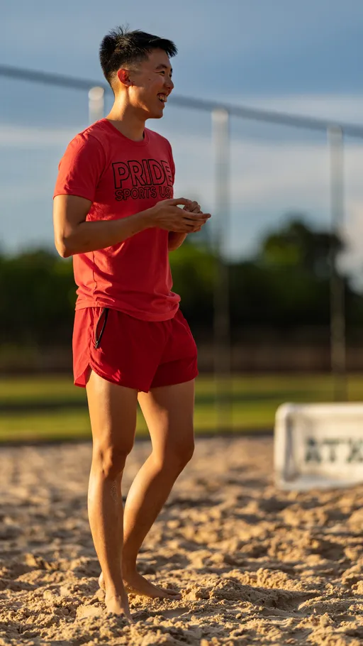 A volleyball player on a sandy outdoor court is smiling and standing barefoot, wearing a red "Pride Sports USA" shirt and red shorts.
