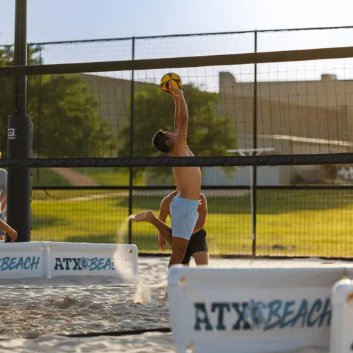 A volleyball player jumps with an outstretched arm to hit the ball over the net on a sand volleyball court, while another player stands behind, both wearing athletic shorts and facing the action. The background shows a sunny outdoor setting with grassy areas and playground equipment.