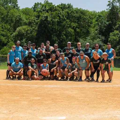 A large group of kickball players, wearing athletic clothing and sunglasses, pose together on a sandy kickball field with trees in the background; several players near the front are holding a red kickball.