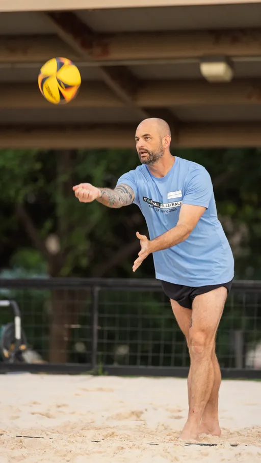A volleyball player wearing a light blue Stonewall Volleyball t-shirt and black shorts is serving a volleyball on a sandy outdoor court, with trees and a fence in the background.