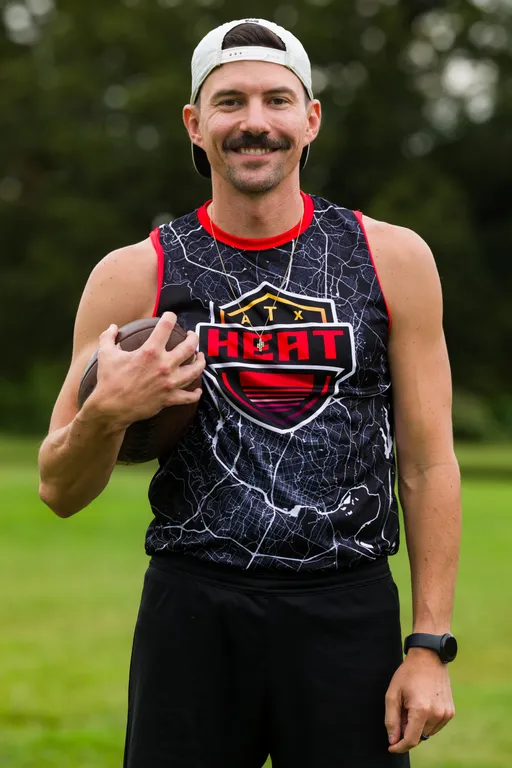 On a grassy field, an ATX Heat (Orange) player preparing for Gay Bowl 2025 holds a football at the chest and faces forward, wearing a black crackle-pattern sleeveless ATX HEAT logo jersey with red trim, a backward light cap, black shorts, a necklace, and a sport watch; trees blur behind.
