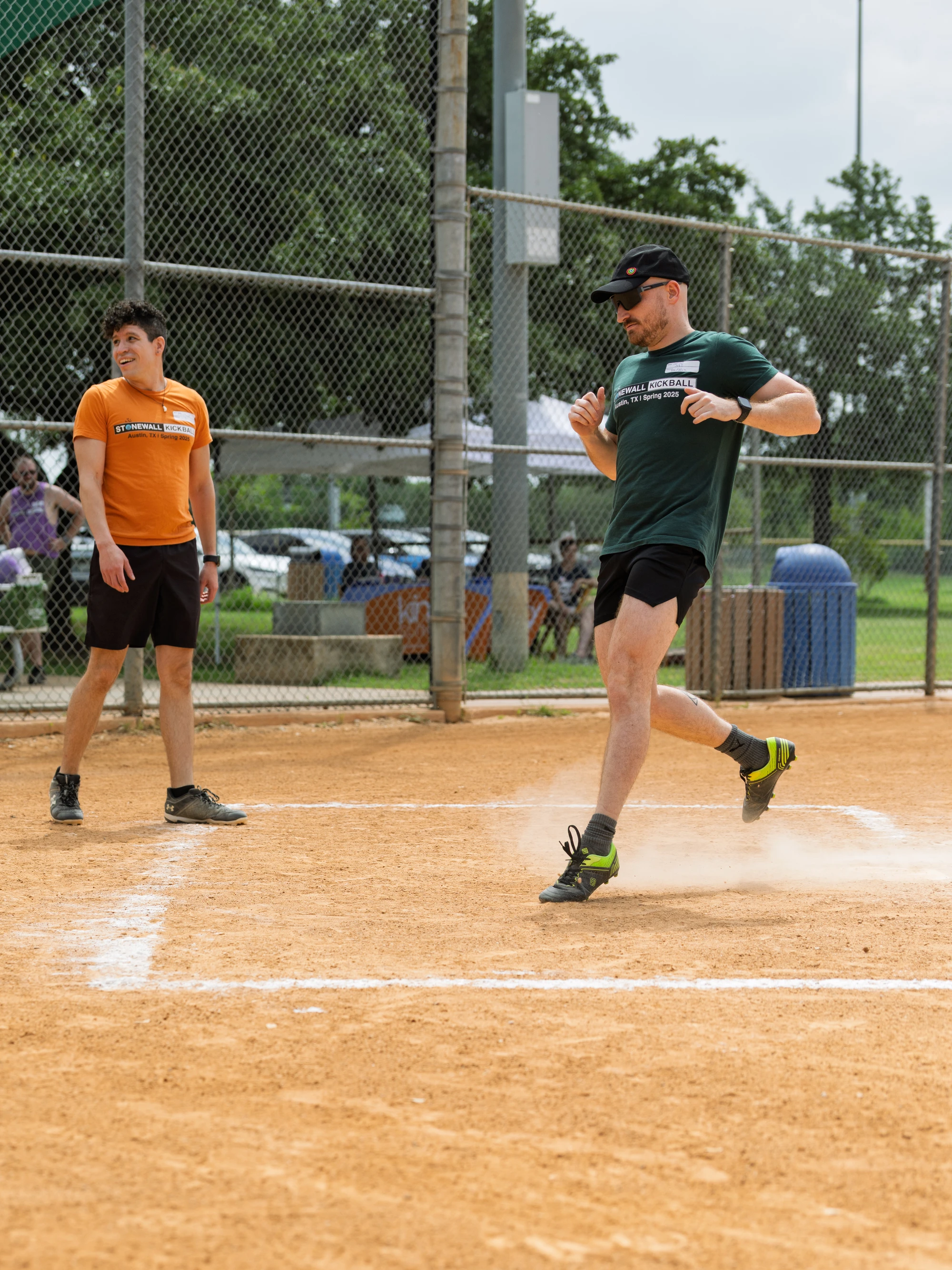 One player in a green shirt, black shorts, and a cap is running across home plate, kicking up dust, while another player in an orange shirt watches from nearby on a kickball field enclosed by a chain-link fence.