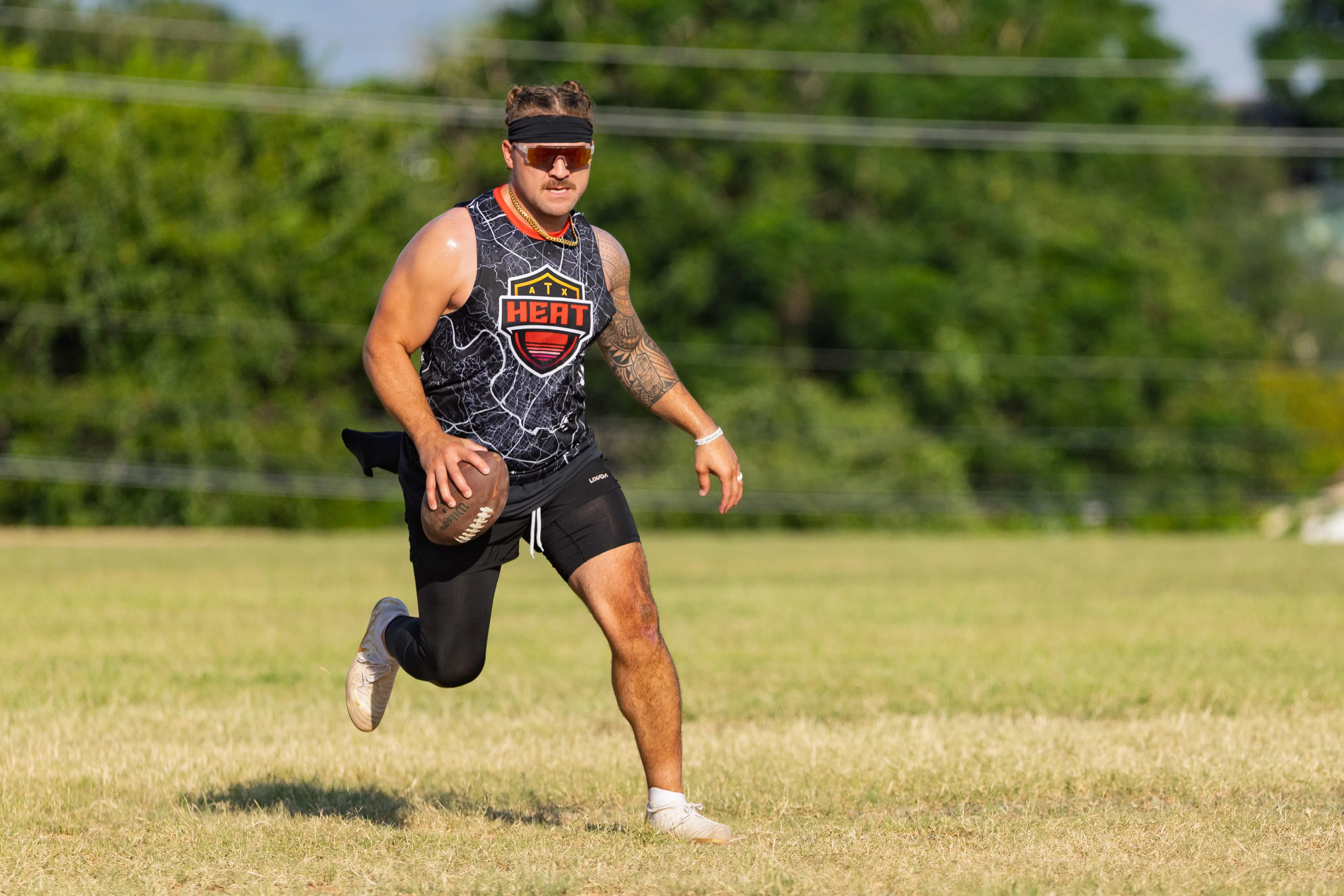 An ATX Heat (Black) player runs across a grassy field during Gay Bowl 2025 practice, cradling a football. The athlete wears a black patterned sleeveless jersey with the ATX Heat crest, black shorts, white shoes, a headband, sport sunglasses, and a flag belt, with trees in the background under bright sun.