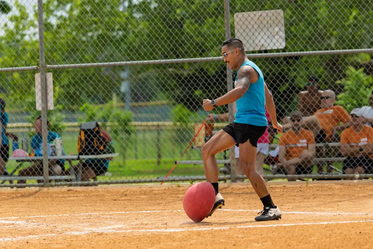 A player in a blue tank top, black shorts, and athletic shoes is about to kick a red kickball on a sandy kickball field, with a crowd of spectators seated behind a chain-link fence in the background.