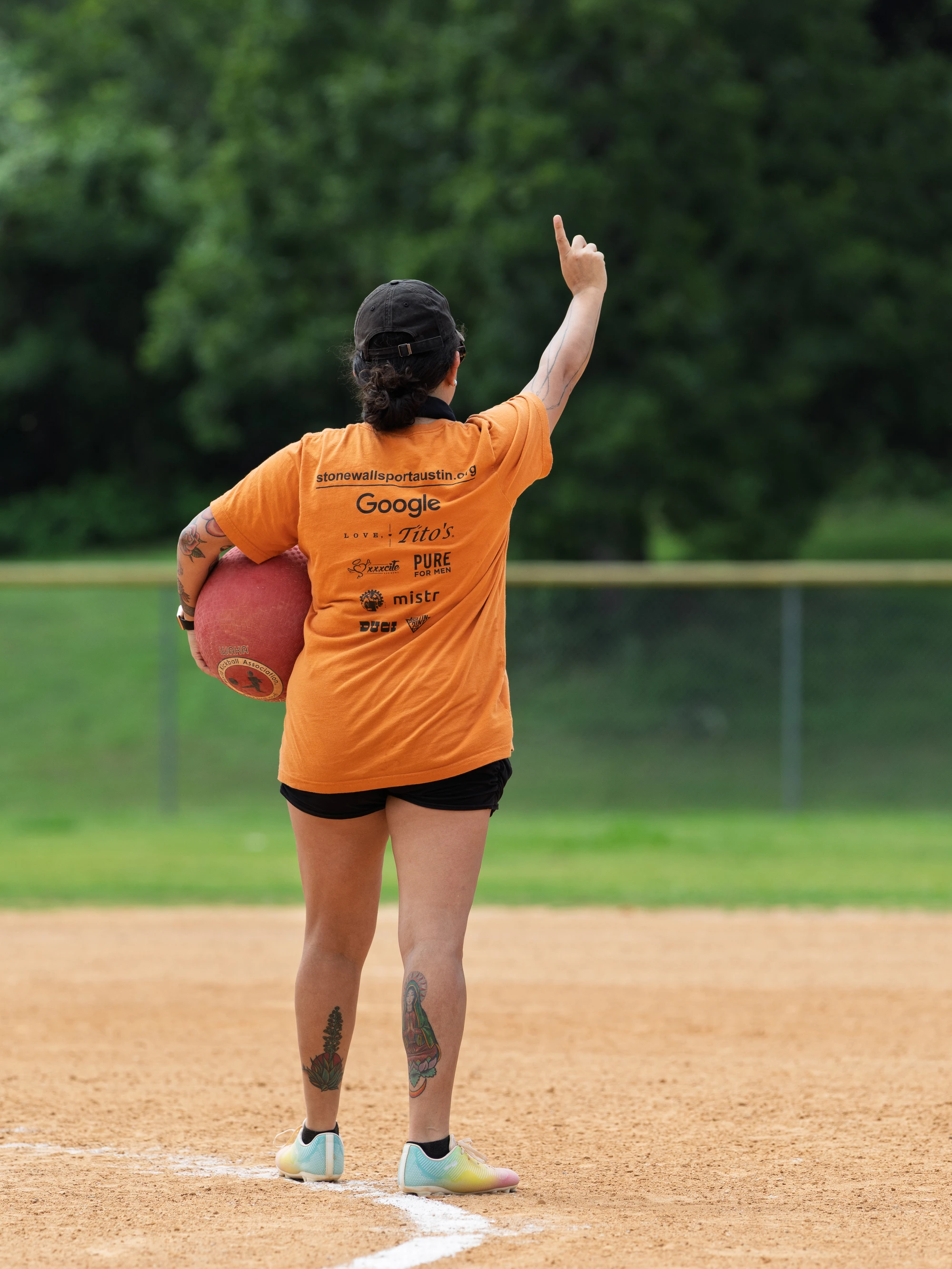 A player wearing an orange shirt and black shorts stands on a kickball field holding a red kickball, raising one arm and pointing upward, with trees and a fence visible in the background.