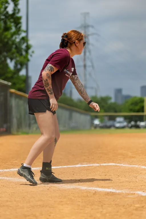 A kickball player wearing a maroon team shirt, black shorts, and athletic shoes is poised and ready near home plate on a dirt kickball field, with trees, a fence, and cars visible in the background.