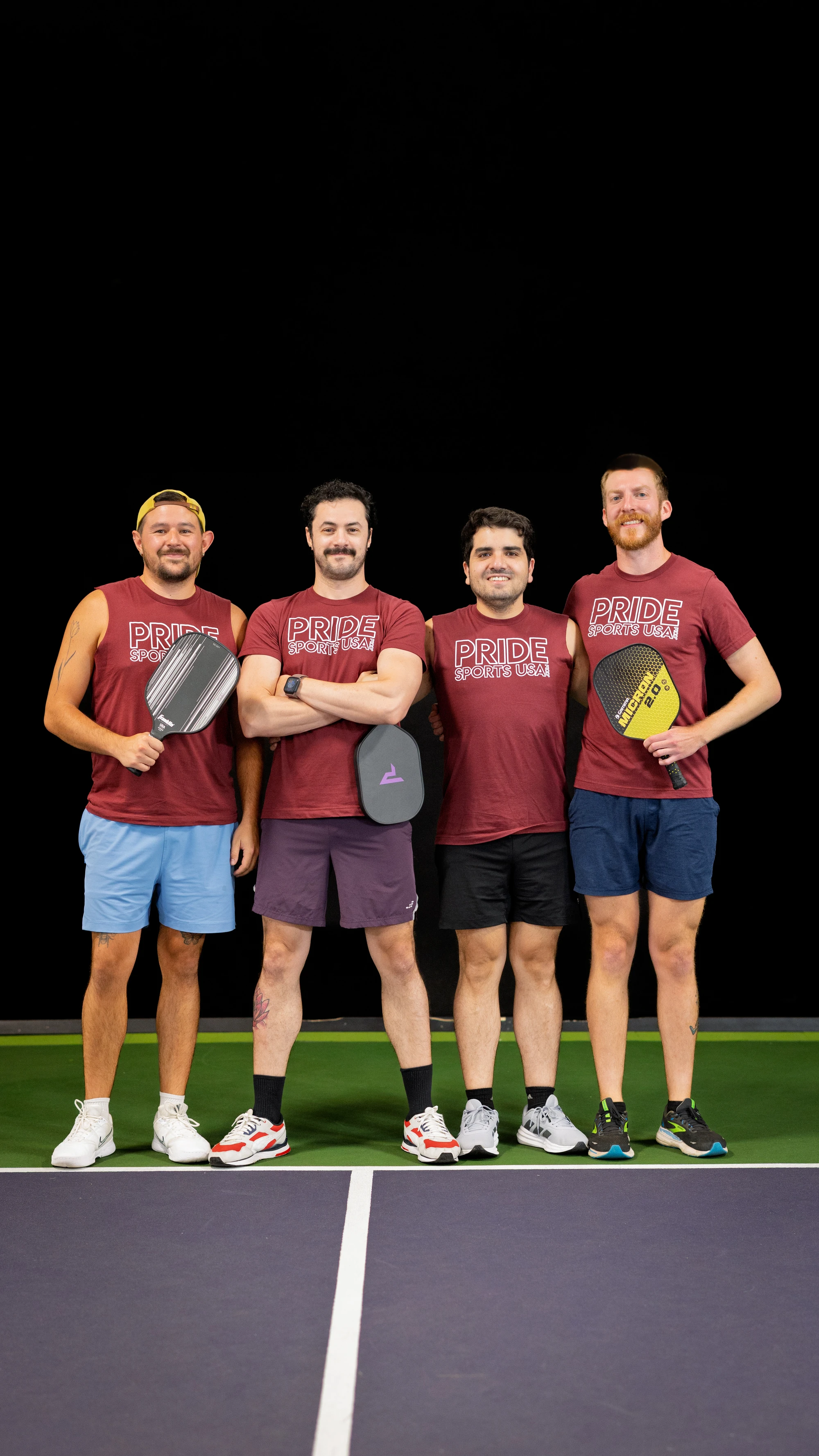 Four pickleball players stand side by side on an indoor pickleball court, each holding a paddle and wearing matching maroon "Pride Sports USA" shirts, athletic shorts, and sneakers.