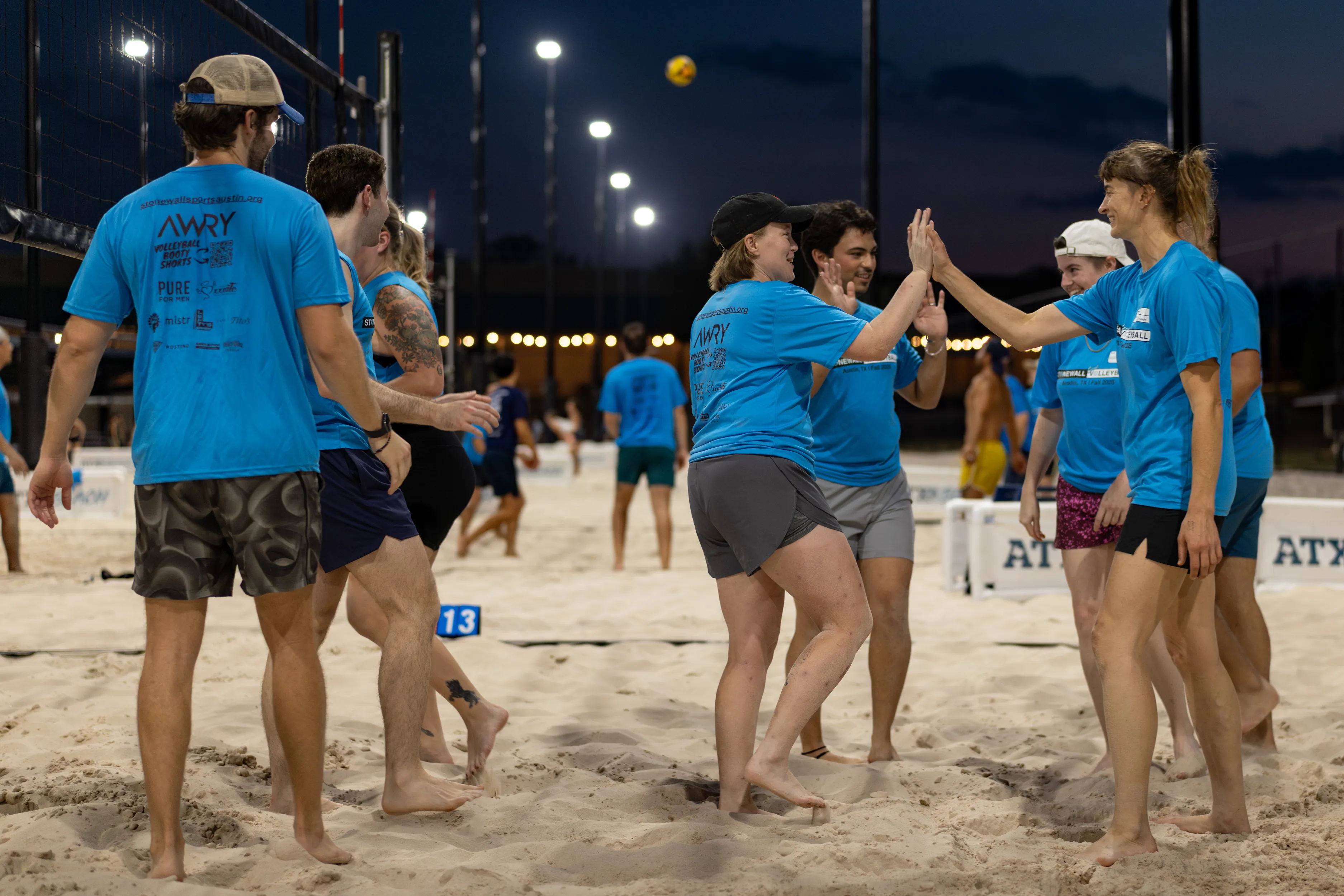 A group of sand volleyball players in matching blue league shirts huddle near the net and exchange high-fives on a lit outdoor court at dusk, wearing shorts and a mix of caps and visors, with other games and a yellow ball visible in the background.