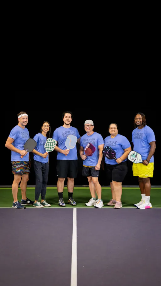 Six pickleball players stand side by side on a pickleball court, each holding a paddle and wearing matching blue "PRIDE" shirts; some are dressed in shorts or athletic wear and are smiling and posing for the photo.