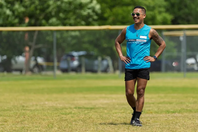 A kickball player stands on the grassy field smiling with hands on hips, wearing a blue Stonewall Kickball Spring 2025 tank top, black shorts, athletic shoes, and sunglasses. Trees, a fence, and parked cars are visible in the background.