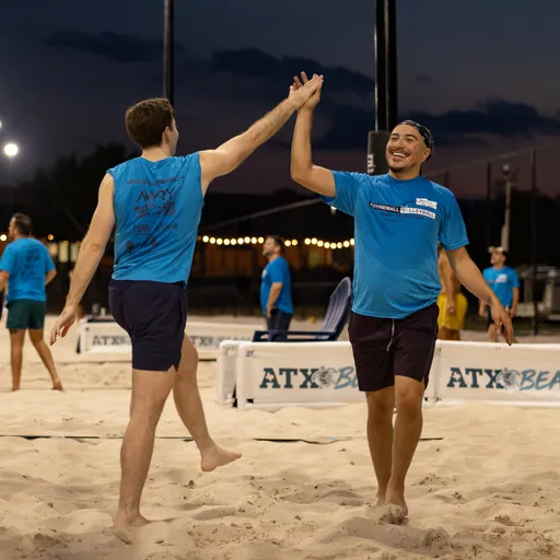 On a lit sand volleyball court at dusk, two players in blue Stonewall league shirts and dark shorts high-five mid-stride, one wearing a headband. Other players in matching shirts stand near multiple nets labeled “ATX,” with string lights and a darkening sky in the background.