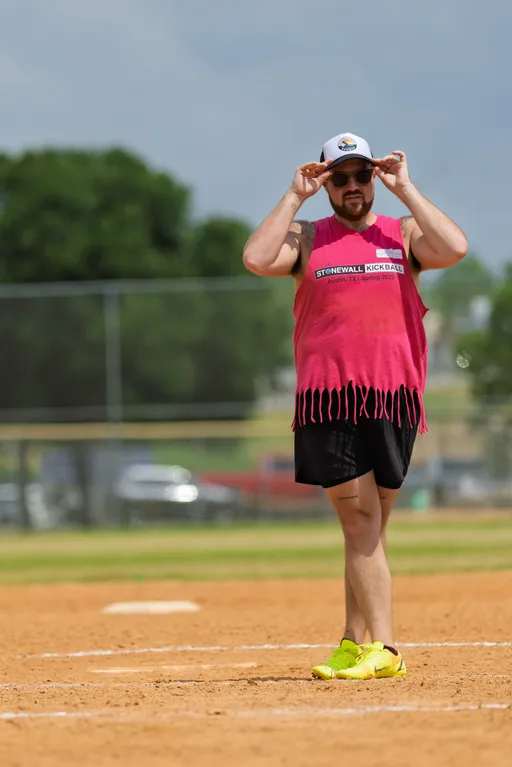 A player stands on a kickball field adjusting their cap and sunglasses, wearing a pink "Stonewall Kickball" fringed tank top, black shorts, and bright yellow athletic shoes.