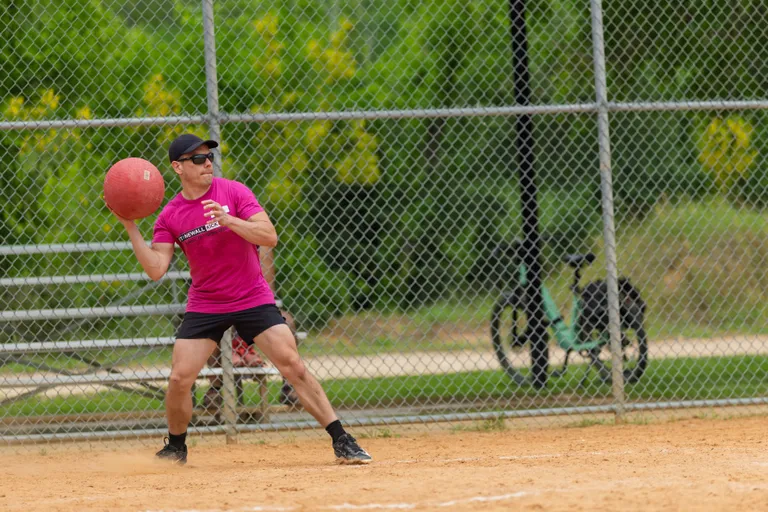 A kickball player wearing a bright pink shirt, black shorts, a black cap, and sunglasses is preparing to throw a red kickball on a sandy field, with a chain-link fence, green trees, and bicycles visible in the background.
