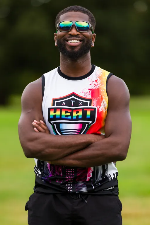 ATX Heat (Orange) player stands on a grassy practice field with arms crossed and smiling, wearing a sleeveless team jersey featuring the “ATX HEAT” shield logo in rainbow colors with an orange side panel and mirrored sunglasses; blurred trees fill the background.