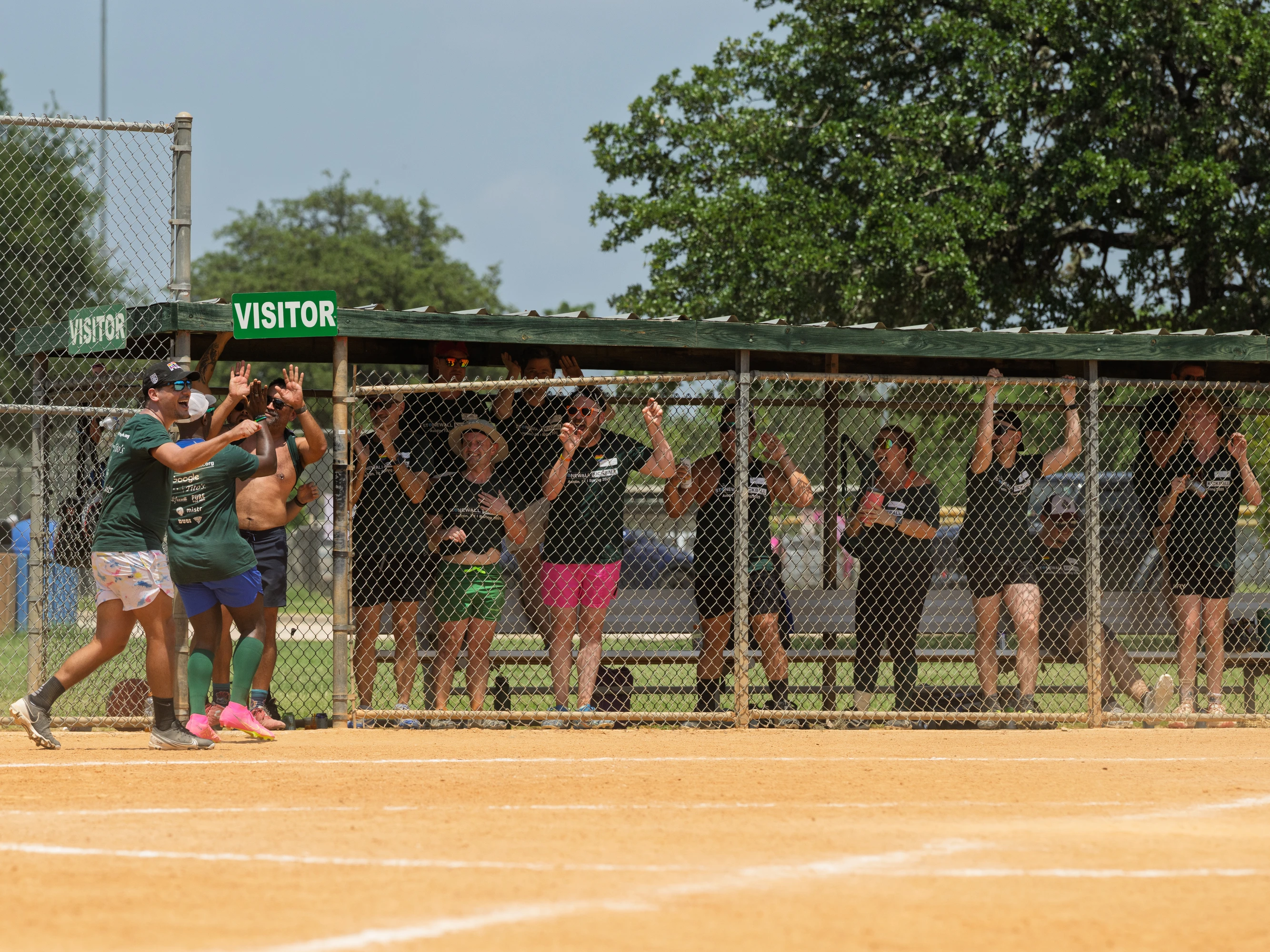 Two kickball players wearing athletic clothing and sneakers celebrate near home plate while members of their team cheer enthusiastically from behind the chain-link fence in the dugout labeled "Visitor"; the scene takes place on a sunny outdoor kickball field with green trees in the background.