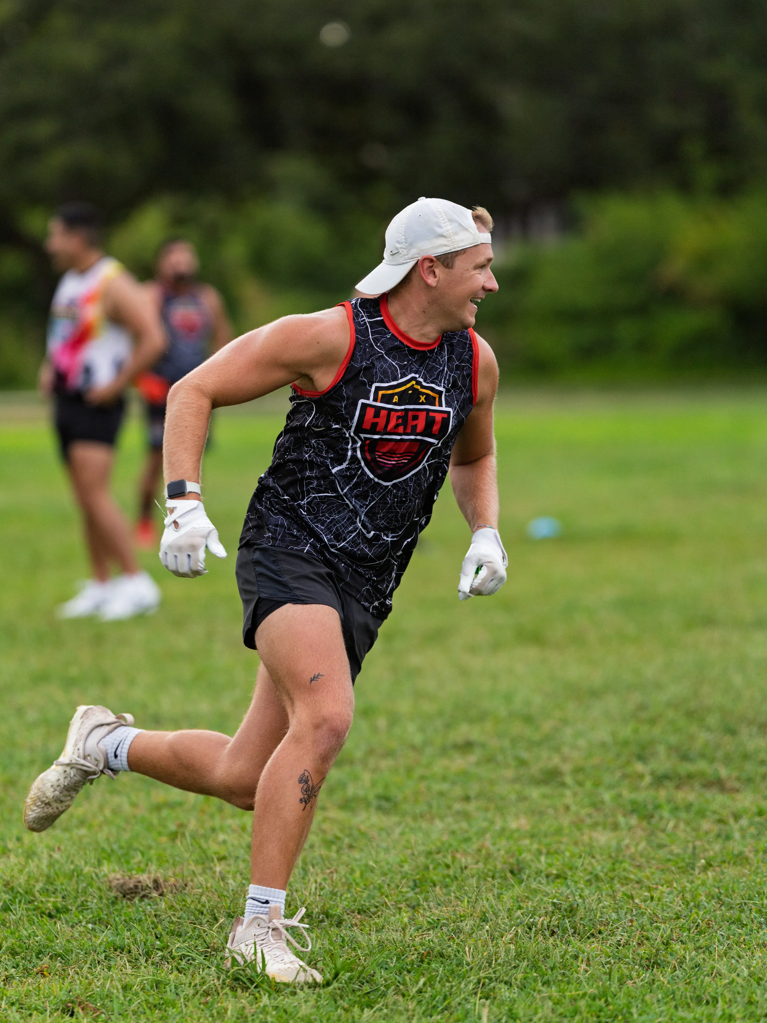A player in a black ATX Heat jersey with red trim, white receiver gloves, black shorts, cleats, and a backward white cap runs across a grassy field during Gay Bowl practice, with teammates and a blue cone blurred in the background.