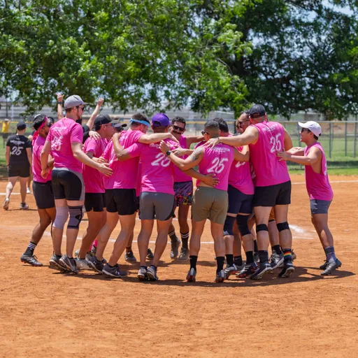 A group of kickball players wearing matching bright pink shirts and athletic gear are huddled together on a dirt kickball field, celebrating or cheering with arms around each other. In the background, a few more people are visible near the edge of the field with green trees behind them.