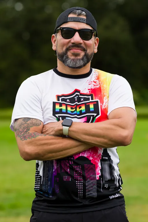 On a grassy practice field, an ATX Heat (Orange) player stands with arms crossed during Gay Bowl 2025 prep, wearing a white-and-orange ATX Heat jersey with a rainbow-shield logo and city-skyline graphic, black shorts, sunglasses, a backward cap, a smartwatch, and a visible forearm tattoo.