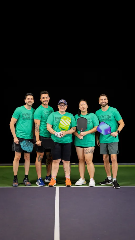 Five pickleball players stand together on an indoor court, each holding a pickleball paddle and wearing matching green "Pride" t-shirts, athletic shorts, and sneakers. The group is positioned behind the net, smiling and appearing ready for a game during the Pride Pickleball Summer 2025 season.