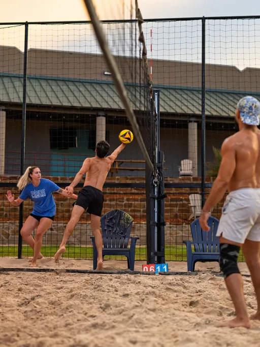 Three athletes play on a sand volleyball court; one player in a blue shirt moves toward the ball while another jumps and reaches up to hit it near the net, and a third player in shorts and a cap observes nearby. The setting includes sand, outdoor seating, and a net with a score marker visible.