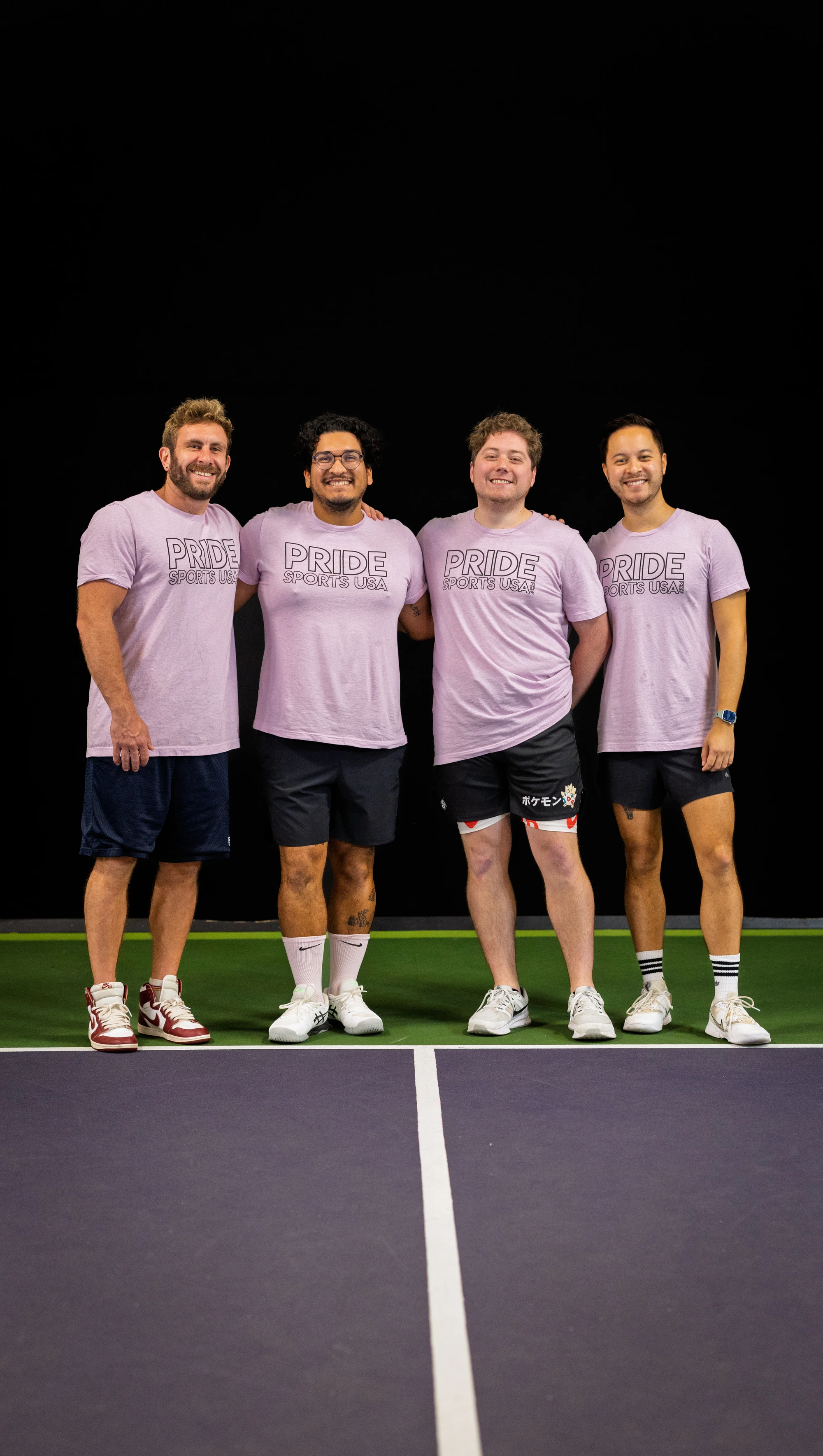 Four pickleball players stand side by side on a court, all wearing matching light purple “Pride Sports USA” t-shirts, athletic shorts, and sneakers, smiling as they pose for a group photo.