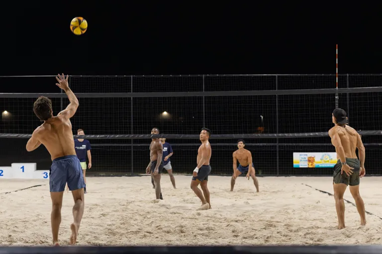 Under stadium lights at a nighttime sand volleyball court, a player on the left tosses a yellow-and-orange ball for a serve while competitors across the net ready their stances; most wear shorts—some shirtless, some in navy tees—with a scoreboard and dark sky in the background.